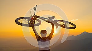 CLOSE UP Man lifts his bicycle above head at sunset after a mountain biking trip