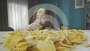 A close-up of a man with long hair and a headband rises in the frame from below. He is drinking beer and eating chips