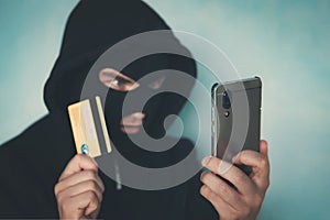 Close up of a man in robbery mask and hood holding the credit card and looking at the smartphone screen. Male criminal arranges a