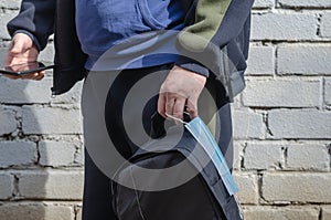 Close-up of a man`s hand holding a backpack by the handle and a medical mask against a white brick wall