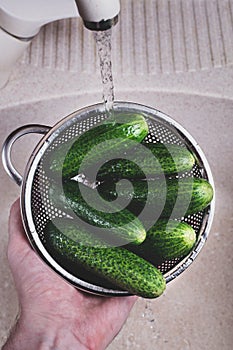 Close-up man`s hand holding cucumbers in a metal colander under running tap water in the sink, health and hygiene washing