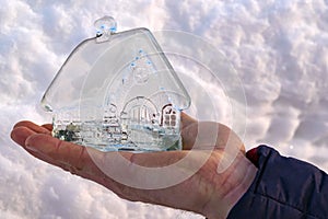 Close-up of man's hand holding small glass, crystal house against background of white snow. Ð¡oncept of dream home