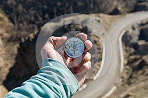 Close-up A man`s hand holds a pocket compass against the backdrop of a mountain road and forest. The concept of outdoor