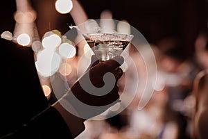 A close up of a man`s hand who is dressed in formal attire and holding a martini glass on party background