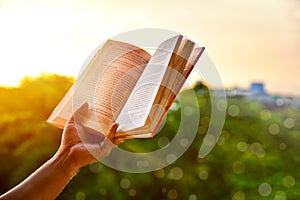 Close-up of man`s hands holding a book by the window for reading against sunset with beautiful nature background.