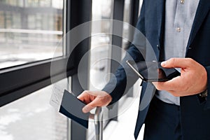 Close up of man`s hands holding phone and passport with ticket. Guy in suit stand at window.