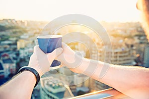 Close up man`s hands holding take away paper cup with morning hot drink - coffee or tea with inspiring view on abstract city back