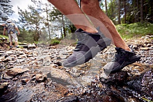 Close-up of man`s legs while crossing a creek during a hiking. Hiking, nature, relationship, together