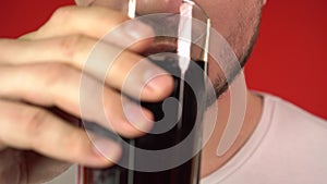 Close-up, a man`s mouth drinks black soda from a glass glass. A man drinks soda. Isolate on a red background, studio