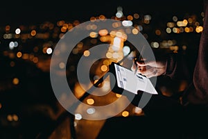 Close-up of man standing on balcony with urban view and using tablet at night