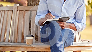 Close Up Of  Man With Takeaway Coffee Sitting On Park Bench Under Tree Writing In Notebook