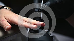 Close-up manicurist's hands in black protective gloves, applying a layer of clear varnish to nail