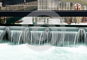 Close up of a manmade concrete weir in a city with a blue river cascading over it and a metal gangway with a rescue ring and equip