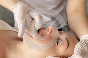 Close-up of masseuse massaging face of young woman lying on couch in salon. Doctor thrusting finger in glove into mouth.