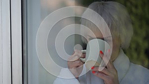 Close-up mature Caucasian woman looking out window drinking morning coffee. Beautiful slim lady in white bathrobe