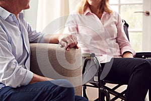 Close Up Of Mature Couple With Woman In Wheelchair Sitting In Lounge At Home Holding Hands Together