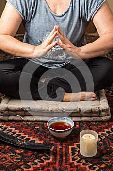 Close-up of mature woman`s body meditating with comfortable clothes, on gray cushion on carpet with incense, cup of tea and candl