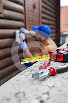 Close up of measuring tape and pliers on concrete fence with professional worker renovating wooden house in the background