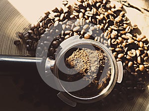 Close up of metal portafilter filled with coffee powder and coffee beans around on wooden table