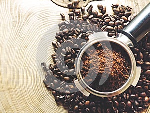 Close up of metal portafilter filled with coffee powder and coffee beans around on wooden table