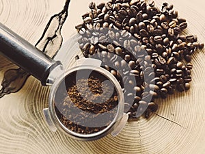 Close up of metal portafilter filled with coffee powder and coffee beans around on wooden table