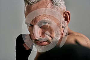 Close up of middle aged athletic man, kickboxer looking agressive while boxing, training in studio over grey background