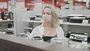 A close-up of a middle-aged blonde woman in a hypermarket walking along the shelves with household electronics, choosing