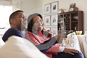 Close up of middle aged couple sitting on the sofa in their living room using remote and watching TV, laughing, close up