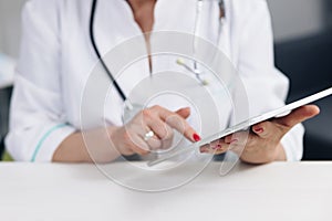 Close up middle aged older female doctor in white uniform holding digital computer tablet in hands, managing patients