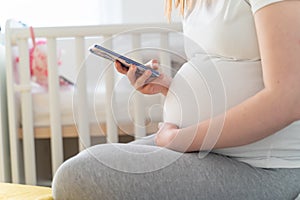 Close up on midsection of unknown caucasian pregnant woman holding mobile phone and her belly during pregnancy while sitting by