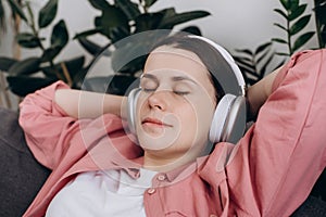 Close up of mindful happy young caucasian female 20s wearing wireless headphones, listening relaxing music, resting on cozy couch