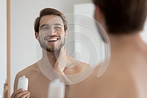 Close up mirror reflection overjoyed young man applying aftershave lotion