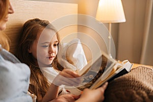 Close-up of mother and happy daughter reading together children book before going to sleep while lying in bed