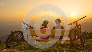CLOSE UP: Mountain bicycle riders sit and observe the sunset from a wooden bench