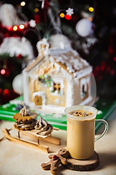 Close-up mug with coffe and milk on a wooden table, gingerbread house and christmas lights and decorations on bokeh