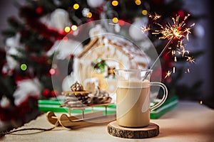 Close-up mug with coffe and milk on a wooden table, gingerbread house and christmas lights and decorations on bokeh. Sparkler