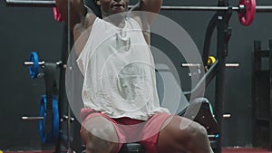 Close up of muscular African American Man sits on bench and lifts up dumbbells in the gym.