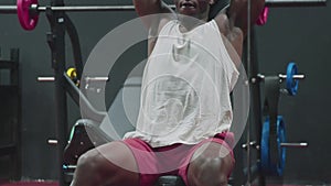 Close up of muscular African American Man sits on bench and lifts up dumbbells in the gym.