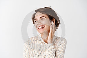 Close-up of natural smiling woman touching earphone in ear and looking at empty space. Girl listening music in