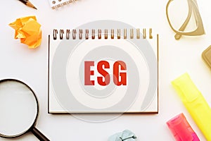 Close-Up of a Notebook With ESG Typography on a White Desk Surrounded by Office Supplies