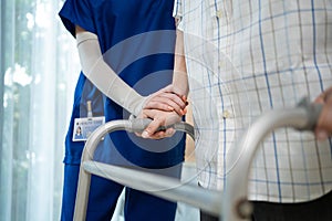 Close up of nurse help disable man walk with walker at nursing home. Caregiver therapist doctor support senior older handicapped