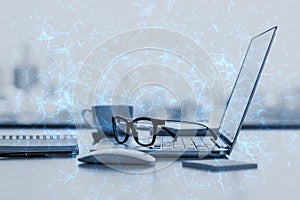 Close up of office desktop with supplies, glasses, coffee cup, laptop and blue polygonal mesh on blurry city view background.