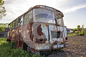 Close-up of old forsaken passenger bus with broken windows rusting in high green weedy grass on edge of plowed brown field on