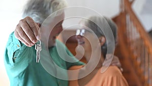 Close up of old and mature woman and man at home holding keys of house to the camera. Portrait of couple of senior smiling and loo