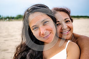 Close up of old mother and pregnant adult daughter are seen hugging and smiling on a beach with white dress