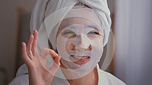 Close up old senior mature Caucasian woman with skincare textile skin mask on female face showing ok good beauty