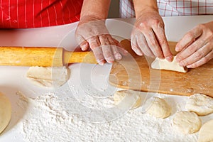Close-up. The older woman in the red kitchen apron rolls out the dough with a rolling pin, the adult daughter sculpts cakes