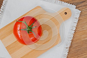 Close-up of one fresh red ripe tomato with water drops on branch, wooden background with rustic chopping board in center