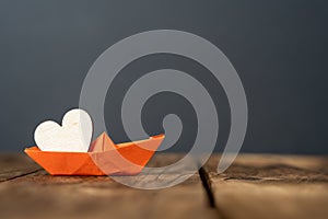 close-up of an orange miniature origami ship with a wooden heart on wooden table.