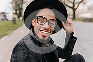 Close-up outdoor portrait of excited young man with brown skin wears glasses. African bearded boy in hat expressing